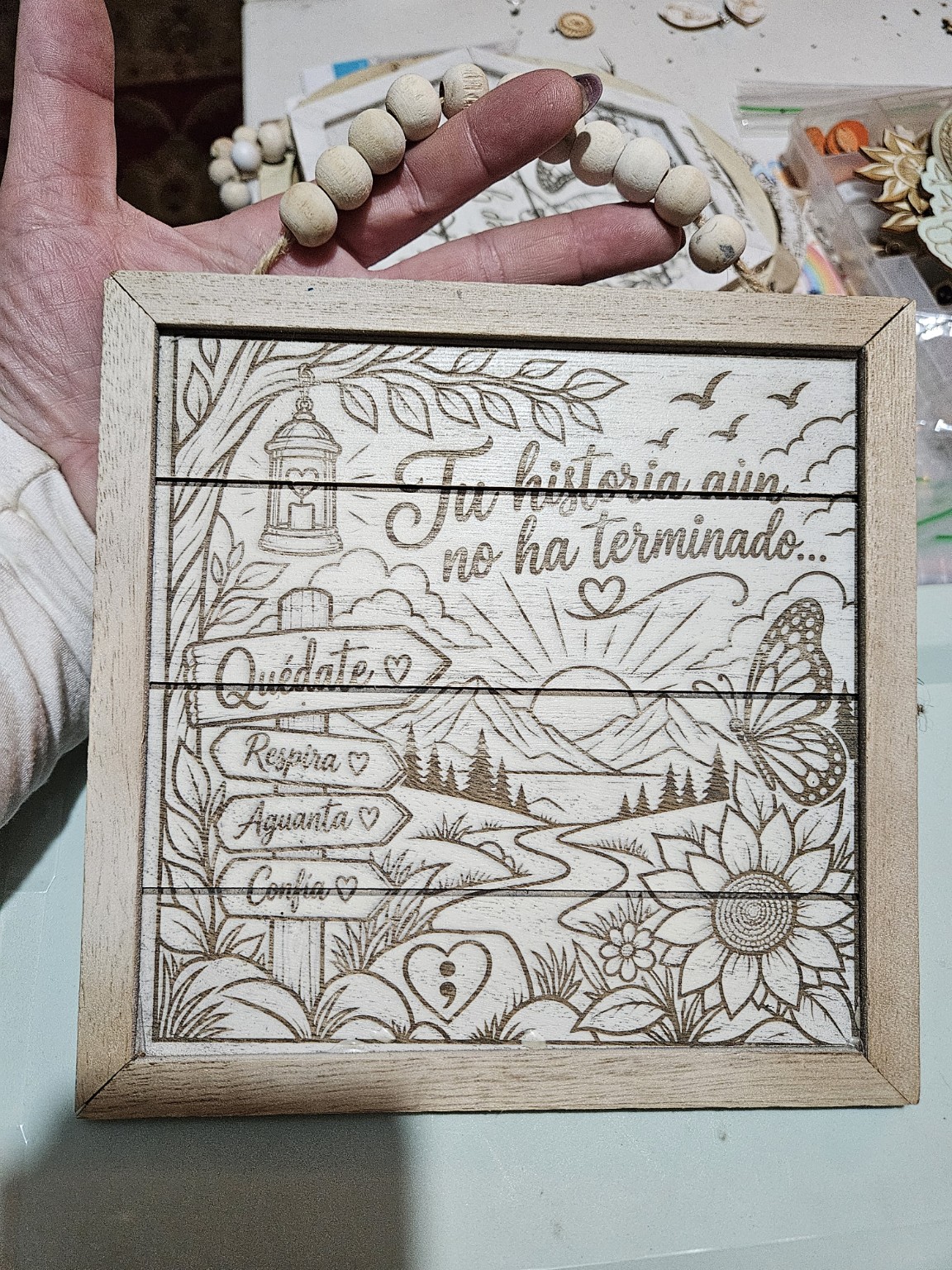 Handmade framed wooden sign in Spanish reading Tu historia aún no ha terminado, with a sunrise, mountains, butterfly, sunflower, and encouraging signposts